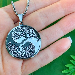 May include: A silver-toned pendant necklace featuring a Yin and Yang design with a tree of life motif. The pendant is circular and hangs from a matching chain. The necklace is displayed against a green background.
