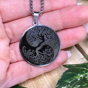 May include: A silver-toned necklace featuring a circular pendant with a black yin-yang design. The pendant showcases a detailed tree of life motif. The necklace is displayed against a backdrop of greenery and a wooden surface.