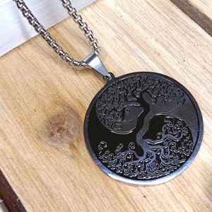 May include: A silver-toned necklace with a circular pendant featuring a black tree of life design. The pendant hangs from a delicate chain, set against a wooden surface with green accents.