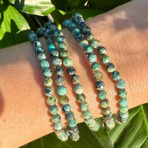 May include: Turquoise beaded bracelets. The bracelets are made of small, round beads in shades of turquoise, green, and gray. The bracelets are stacked on top of each other. The background is a green leaf.