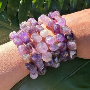 May include: A set of five amethyst gemstone bracelets, each with a different shade of purple. The bracelets are made of smooth, rounded stones and are stacked on a person's wrist.