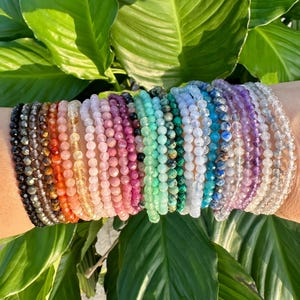 Pode incluir: Uma coleção de pulseiras de contas em várias cores, incluindo preto, laranja, rosa, verde e roxo. As pulseiras são exibidas em um fundo de folhas verdes exuberantes, criando uma estética vibrante e natural.