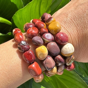 Op de afbeelding: Een set van drie armbanden gemaakt met kleurrijke, gepolijste, natuurlijke stenen kralen. De kralen zijn rood, oranje, geel, bruin en wit. De armbanden zijn gestapeld op de pols van een persoon.