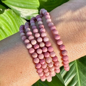 Pode incluir: Várias pulseiras de contas rosa. As contas são pequenas, redondas e de uma cor rosa suave. As pulseiras estão enroladas em um pulso, com folhas verdes ao fundo.