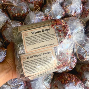 May include: A close-up of a bundle of white sage and lavender smudge sticks wrapped in clear plastic with a brown twine bow. The bundle is labelled "White Sage & Lavender" and includes information about the plant's history and uses. The label also includes the website address "www.MayanRoseShop.Etsy.com".