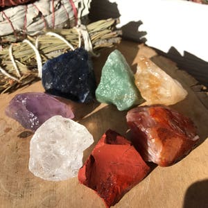 May include: Seven chakra crystals, including amethyst, clear quartz, green aventurine, citrine, carnelian, red jasper, and sodalite, arranged in a circle on a wooden surface.