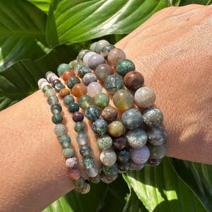 May include: A stack of seven bracelets made of green, brown, and white agate beads. The bracelets are stacked on a person's wrist.