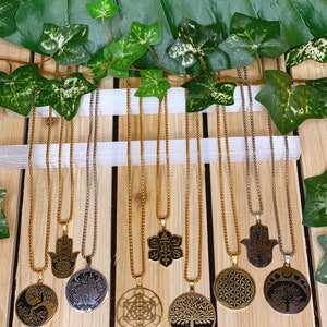May include: A collection of gold and silver-toned necklaces with various sacred symbols, including a tree of life, lotus flower, and Hamsa hand. The necklaces hang against a wooden background, with the text "EXPLORE THE SACRED SYMBOLS NECKLACE COLLECTION" at the top.