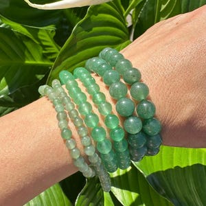 May include: Six green beaded bracelets in varying shades of green are stacked on a wrist. The bracelets feature round beads of different sizes. The background includes green leaves and a white flower.