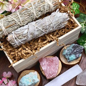 May include: A wooden box filled with sage smudge sticks, crystals, and a selenite wand. The box is surrounded by pink flowers and green leaves.