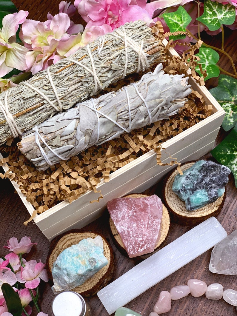 May include: A wooden box filled with sage smudge sticks, crystals, and a selenite wand. The box is surrounded by pink flowers and green leaves.