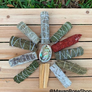 May include: A collection of various smudge sticks, including white sage, palo santo, cedar, and red sage, arranged in a circle on a wooden surface. A seashell is in the centre of the circle. The text "@MayanRoseShop" is at the bottom of the image.