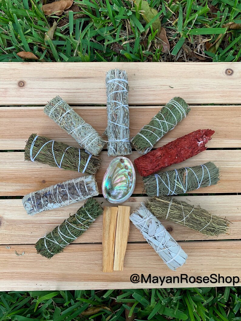 May include: A collection of various smudge sticks, including white sage, palo santo, cedar, and red sage, arranged in a circle on a wooden surface. A seashell is in the centre of the circle. The text "@MayanRoseShop" is at the bottom of the image.