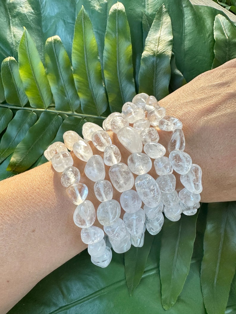 May include: A stack of clear quartz crystal bracelets with a smooth, rounded pebble shape. The bracelets are stacked on a person's wrist.