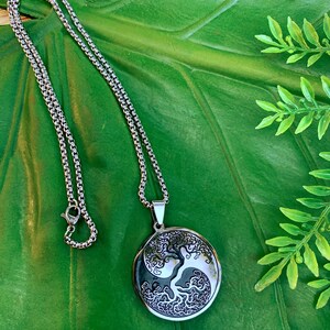 May include: Silver-toned necklace with a round pendant featuring a tree design within a yin-yang symbol. The pendant hangs from a chain with a lobster clasp. The necklace rests on a large green leaf.