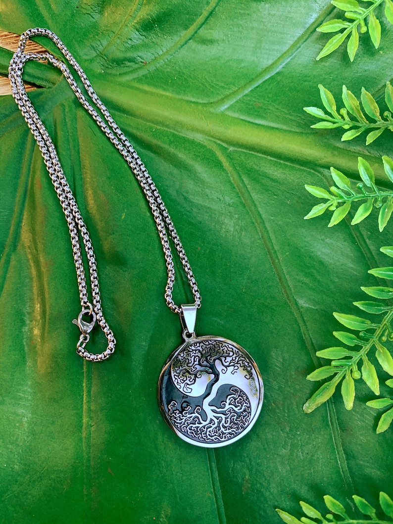 May include: Silver-toned necklace with a round pendant featuring a tree design within a yin-yang symbol. The pendant hangs from a chain with a lobster clasp. The necklace rests on a large green leaf.