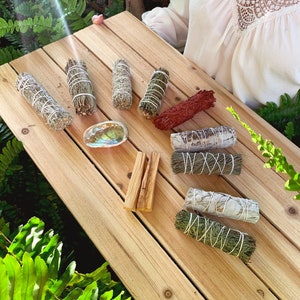 May include: A selection of smudge sticks on a wooden surface. The smudge sticks are made from different herbs and are tied with twine. There are also two sticks of palo santo wood.