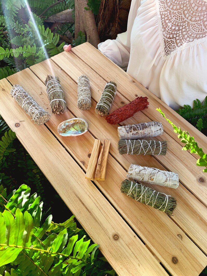 May include: A selection of smudge sticks on a wooden surface. The smudge sticks are made from different herbs and are tied with twine. There are also two sticks of palo santo wood.