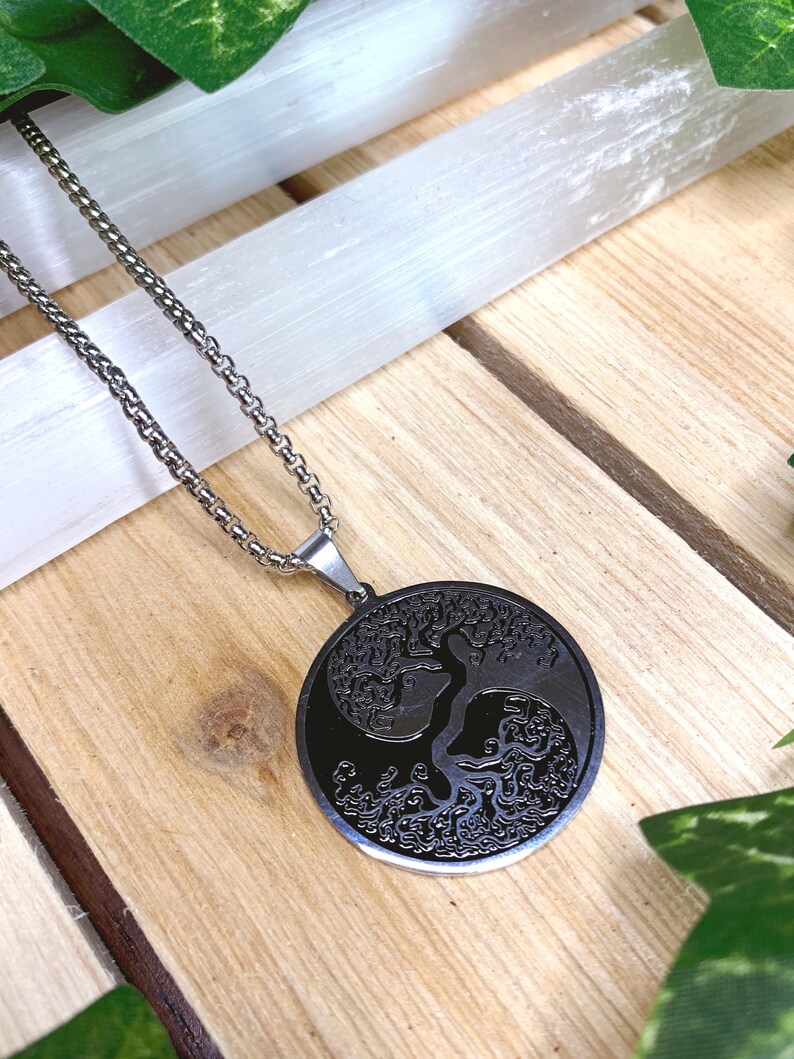 May include: A silver-toned necklace with a circular pendant featuring a black tree of life design. The pendant hangs from a delicate chain, set against a wooden surface with green accents.