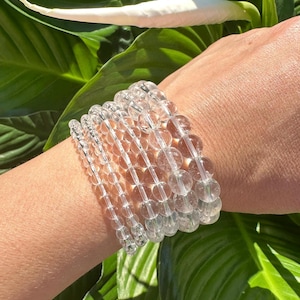 Könnte beinhalten: Ein Stapel aus Armbändern aus klaren Quarzkristallperlen. Die Armbänder bestehen aus runden, glatten Perlen und sind auf dem Handgelenk einer Person gestapelt.