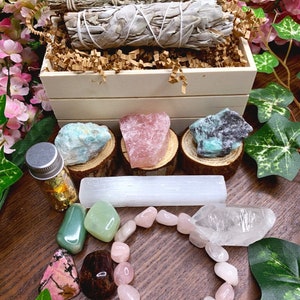 May include: A wooden box filled with crystals, sage smudge sticks, and a selenite wand. The box is surrounded by green leaves and pink flowers. There is also a small bottle of gold leaf and a pink crystal bracelet.