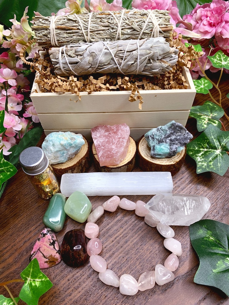 May include: A wooden box filled with crystals, sage smudge sticks, and a selenite wand. The box is surrounded by green leaves and pink flowers. There is also a small bottle of gold leaf and a pink crystal bracelet.