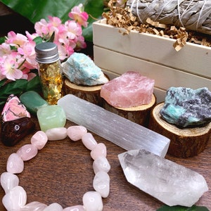 May include: A wooden box filled with crystals, including rose quartz, amethyst, and selenite. There is also a small bottle of gold leaf and a sage smudge stick. The crystals are arranged on a wooden surface with green leaves.