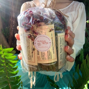 May include: A close-up of a bundle of dried herbs wrapped in clear plastic. The bundle includes white sage, cedar, and rosemary. The label reads "White Sage & Cedar" and "Rosemary". The label also includes the text "Mayan Rose Shop" and "Thank you for your purchase".
