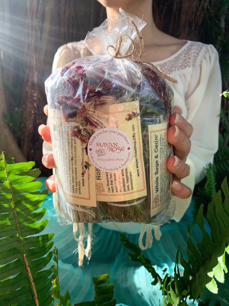 May include: A close-up of a bundle of dried herbs wrapped in clear plastic. The bundle includes white sage, cedar, and rosemary. The label reads "White Sage & Cedar" and "Rosemary". The label also includes the text "Mayan Rose Shop" and "Thank you for your purchase".