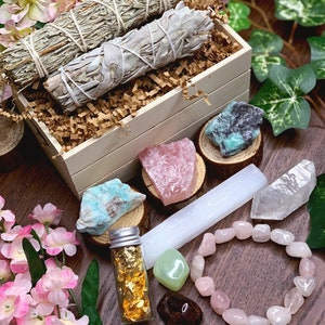 May include: A wooden box filled with sage smudge sticks, crystals, and a small bottle of gold leaf. The box is surrounded by pink flowers and green leaves.