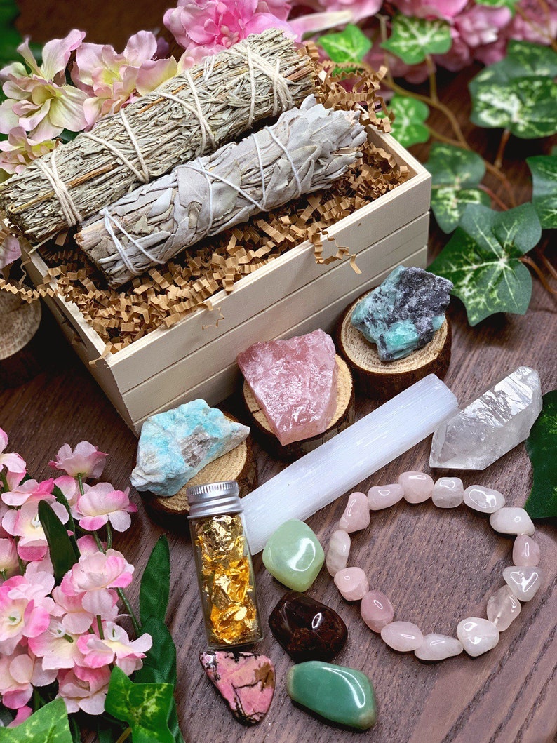 May include: A wooden box filled with sage smudge sticks, crystals, and a small bottle of gold leaf. The box is surrounded by pink flowers and green leaves.