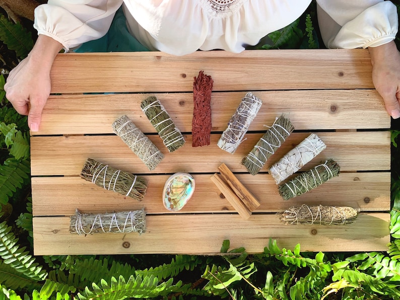 May include: A selection of smudge sticks arranged in a semi-circle on a wooden surface. The sticks are made from different herbs and woods, including sage, palo santo, and white sage. There is also a small abalone shell and two pieces of wood in the centre of the arrangement.