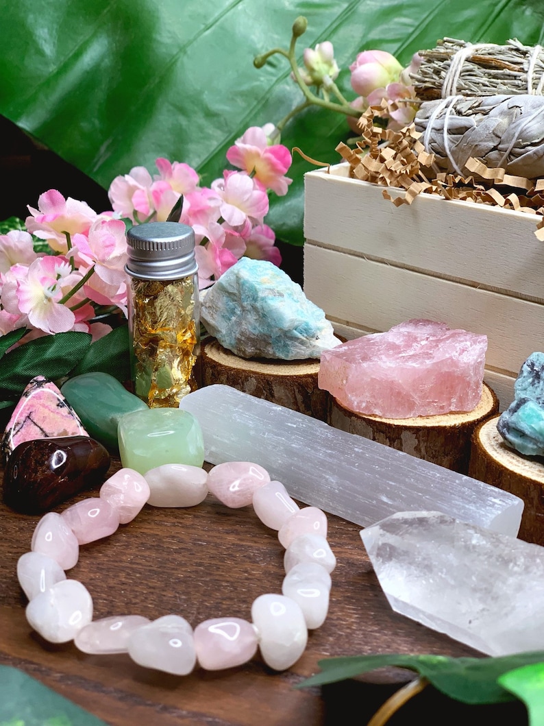 May include: A collection of crystals and stones, including rose quartz, selenite, and amethyst, arranged on a wooden surface. There is also a small bottle of gold leaf and a pink flower bouquet.