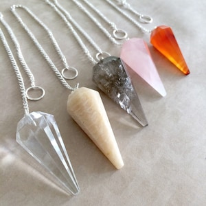May include: Six crystal pendulums with silver chains hanging from a light brown surface. The pendulums are made of clear quartz, rose quartz, smoky quartz, carnelian, and a light beige stone.