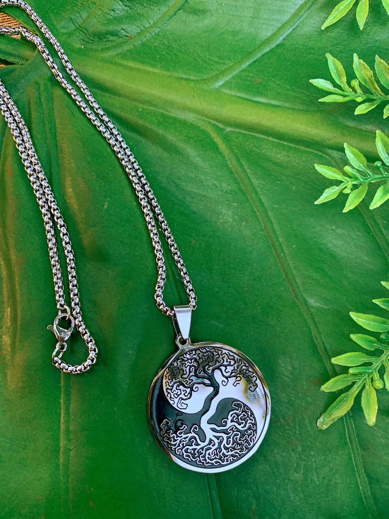 May include: A silver-colored necklace with a round pendant featuring a tree design within a yin-yang symbol. The necklace chain is a box chain style. The pendant is approximately 1.5 inches in diameter. The background is a large green leaf.