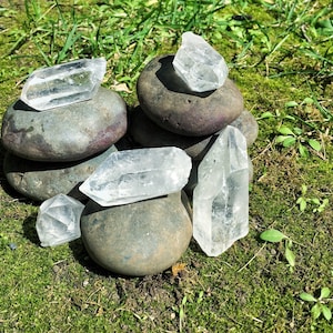 May include: A group of clear quartz crystals arranged on top of a stack of gray stones. The crystals are various shapes and sizes, and the stones are smooth and rounded.