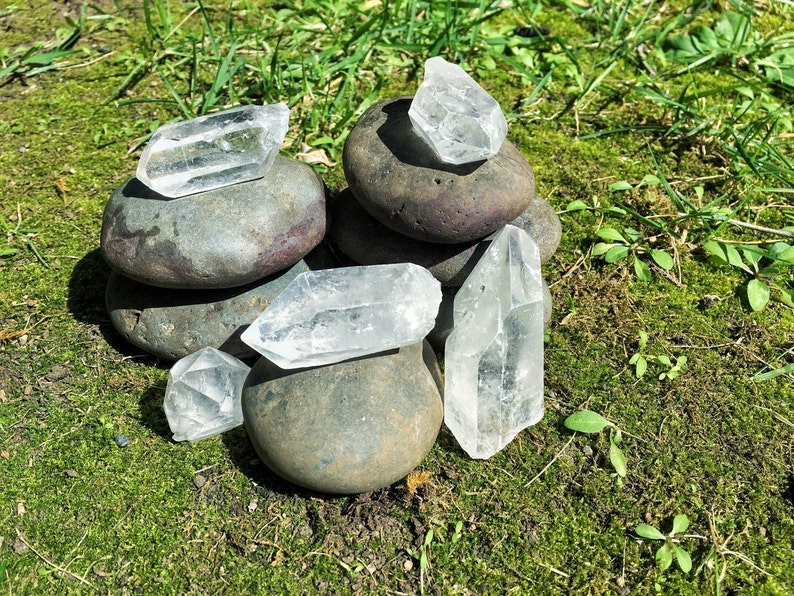 May include: A group of clear quartz crystals arranged on top of a stack of gray stones. The crystals are various shapes and sizes, and the stones are smooth and rounded.