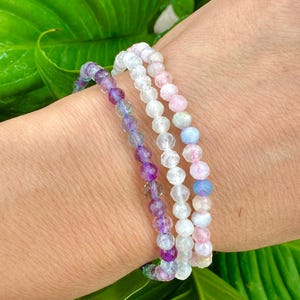 May include: Three beaded bracelets in shades of purple, white, and pink are stacked on a wrist. The bracelets are made of small, faceted beads. The background is a blurred green leafy plant.