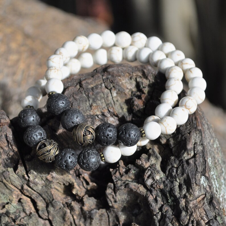 May include: Two bracelets, one with white beads and the other with black lava rock beads and gold accents. The bracelets are layered on a piece of wood.