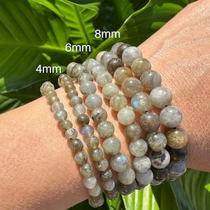 May include: A selection of gemstone bead bracelets in various sizes (4mm, 6mm, and 8mm) are shown. The beads are grey and brown. The bracelets are displayed on a wrist, with green leaves visible in the background.
