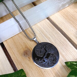 May include: A silver-toned necklace with a circular pendant featuring a tree of life design. The pendant is black with intricate details, and the chain is a classic link style. The necklace rests on a wooden surface.