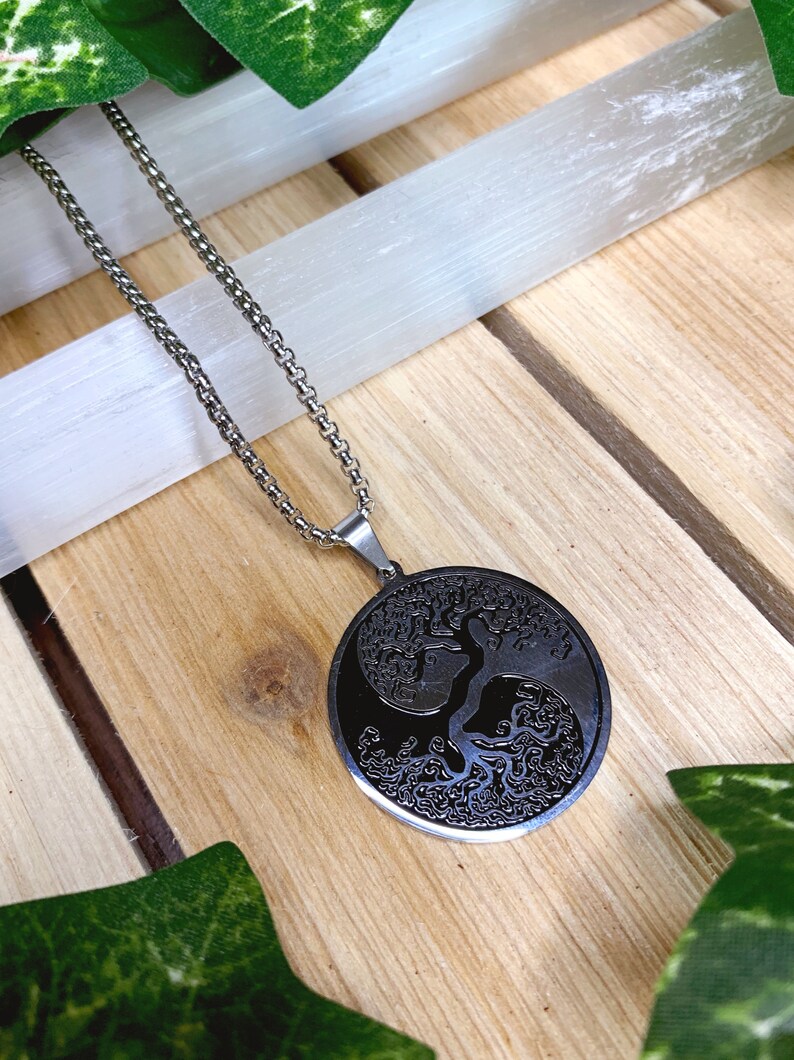 May include: A silver-toned necklace with a circular pendant featuring a tree of life design. The pendant is black with intricate details, and the chain is a classic link style. The necklace rests on a wooden surface.