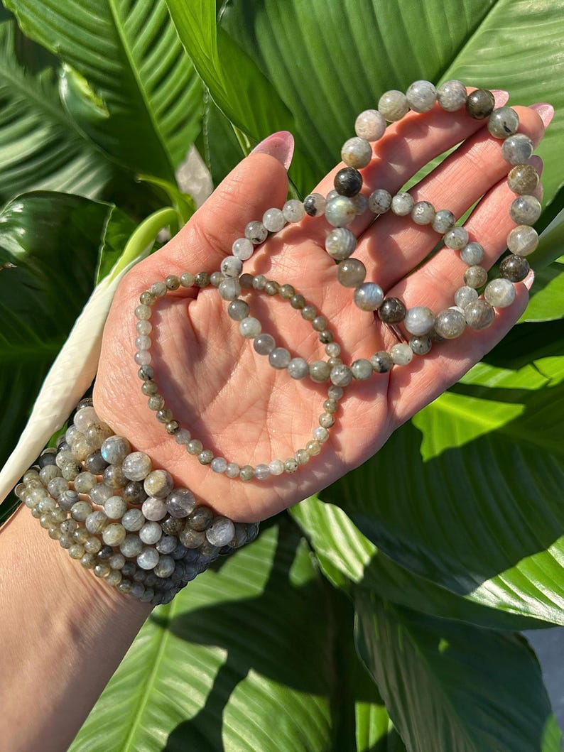 May include: A collection of labradorite bead bracelets in different sizes. The bracelets are made of grey, green, and iridescent beads. The bracelets are displayed against a background of green leaves.
