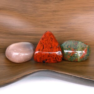 Puede incluir: Tres piedras pulidas, una roja, una verde con motas rojas y blancas, y una rosa p&aacute;lida, sobre una superficie de madera marr&oacute;n.