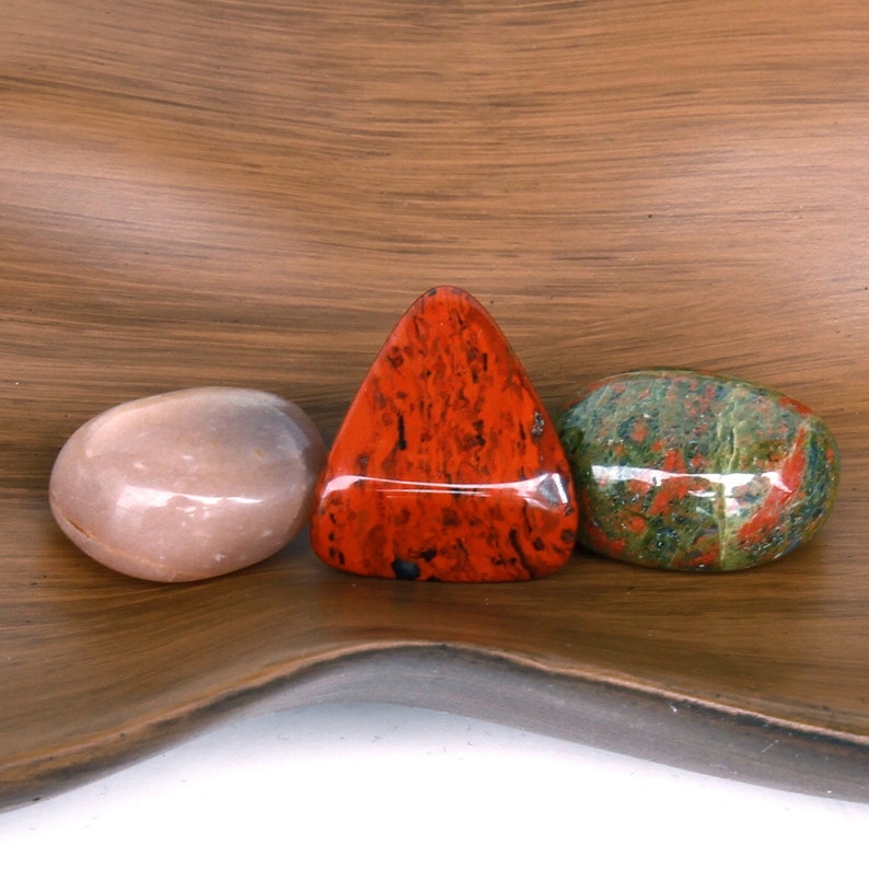 Puede incluir: Tres piedras pulidas, una roja, una verde con motas rojas y blancas, y una rosa p&aacute;lida, sobre una superficie de madera marr&oacute;n.