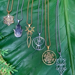 May include: An assortment of pendant necklaces with various spiritual and geometric designs. The necklaces feature gold and silver-toned chains and pendants, including a lotus flower, Hamsa hand, and Metatron's Cube. The pendants are displayed on a large green leaf.