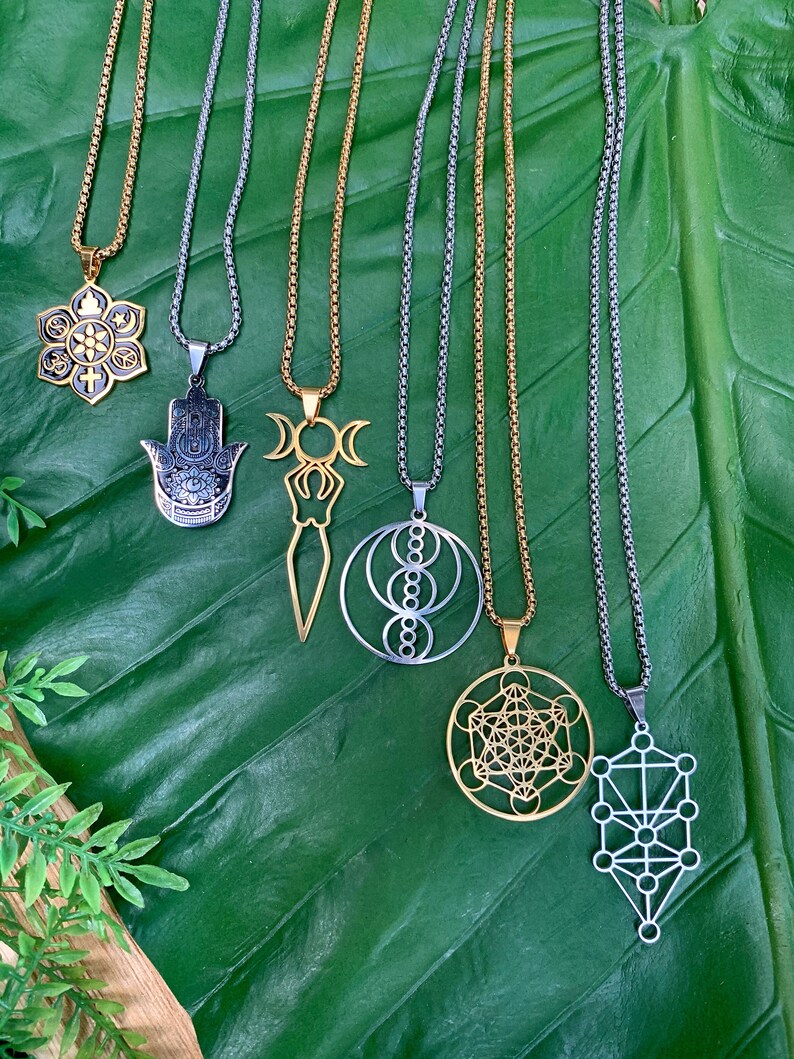 May include: An assortment of pendant necklaces with various spiritual and geometric designs. The necklaces feature gold and silver-toned chains and pendants, including a lotus flower, Hamsa hand, and Metatron's Cube. The pendants are displayed on a large green leaf.