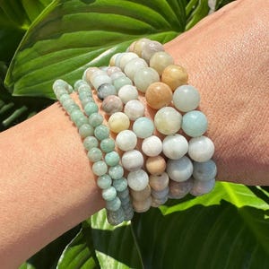 May include: A stack of bracelets made with green, white, and brown gemstone beads. The bracelets are worn on a person's wrist.