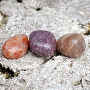 May include: Three smooth, polished stones in a row on a gray, textured surface. The stones are orange, purple, and pink.