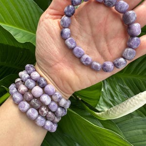 Puede incluir: Un conjunto de cuatro pulseras de piedras preciosas moradas con cuentas lisas y redondeadas. Las pulseras est&aacute;n apiladas en la mu&ntilde;eca de una persona.